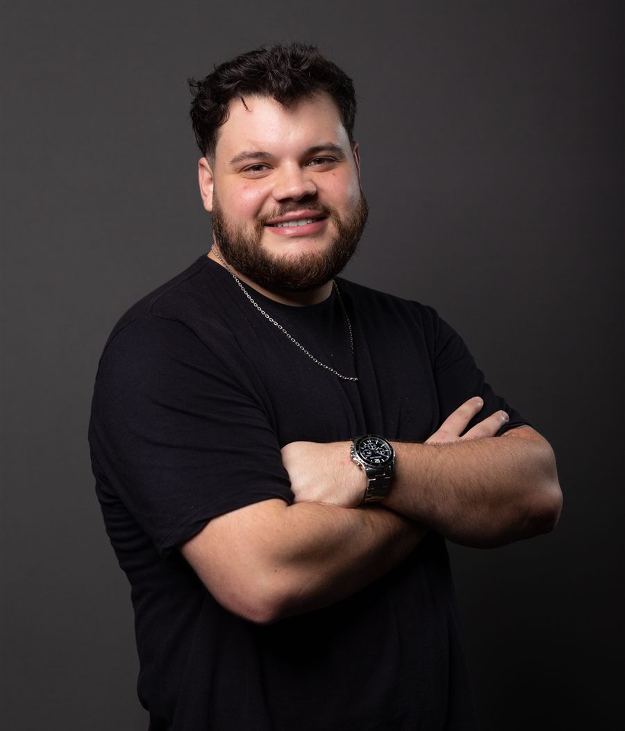 Bruno Holzmann, de camiseta preta e braços cruzados, sorrindo, em fundo cinza
